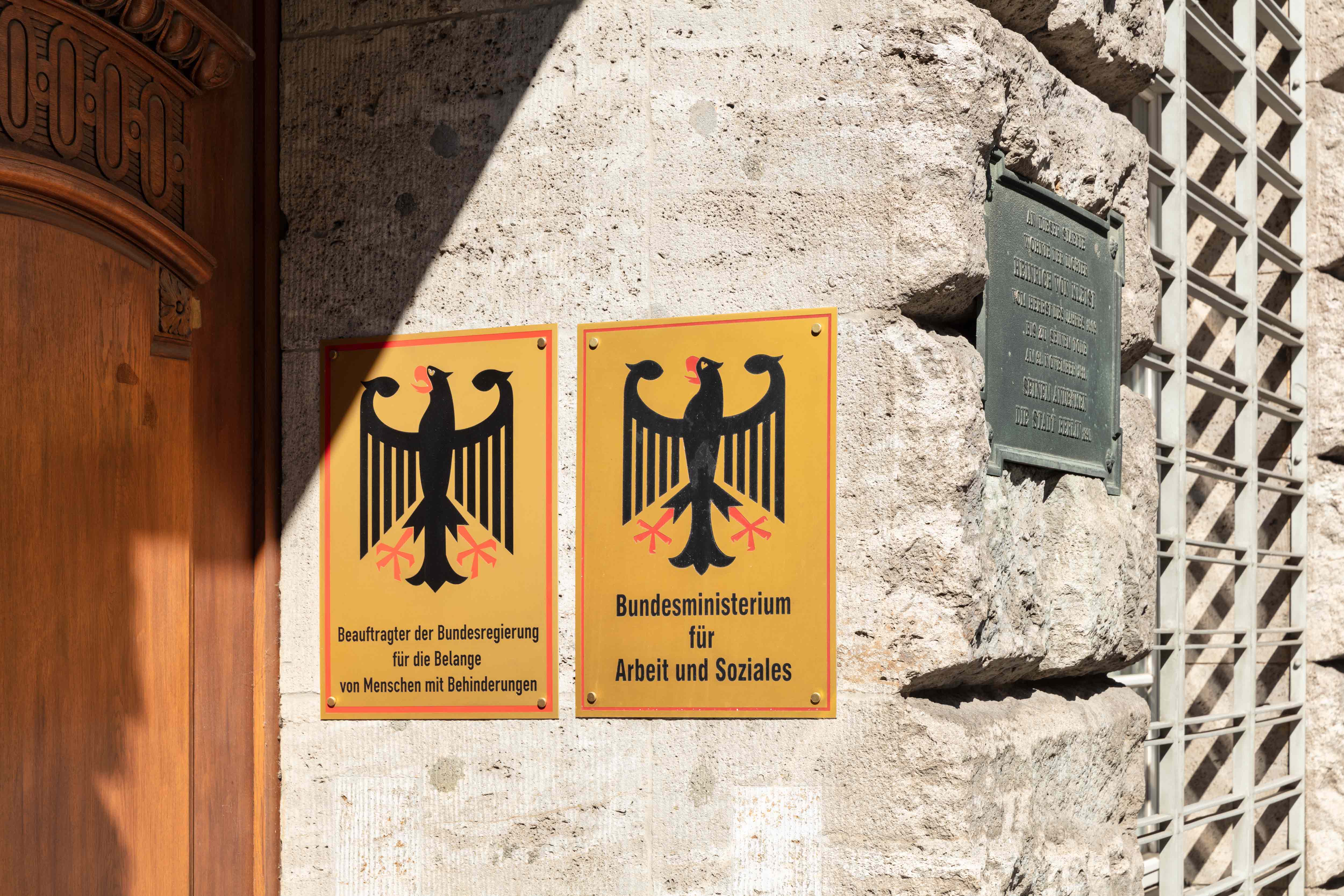Eingang des Kleisthauses: Links eine große braune Holztür. Rechts eine Steinwand mit zwei Schildern: Beauftragter der Bundesregierung für die Belange von Menschen mit Behinderungen und Bundesministerium für Arbeit und Soziales (Quelle: BMAS/Pinjo) Eingang des Kleisthauses: Links eine große braune Holztür. Rechts eine Steinwand mit zwei Schildern: Beauftragter der Bundesregierung für die Belange von Menschen mit Behinderungen und Bundesministerium für Arbeit und Soziales