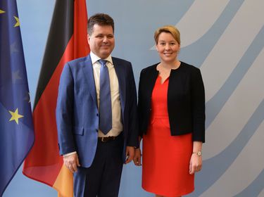 Jürgen Dusel und Franziska Giffey stehen vor einer blauen Wand, rechts ist die „Adlerschwinge“ des Logos der Bundesregierung zu sehen, links stehen zwei Flaggen: Die europäische und die deutsche Flagge. Dusel und Giffey lächeln in die Kamera.