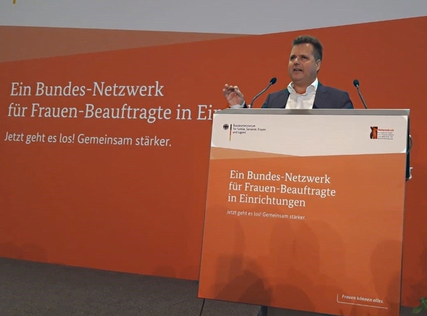 Jürgen Dusel steht an einem Rednerpult und spricht. Hinter ihm auf der Rückwand und auch auf dem Rednerpult steht: „Ein Bundes-Netzwerk für Frauen-Beauftragte in Einrichtungen“ und „Jetzt geht es los! Gemeinsam stärker. Frauen können alles“.