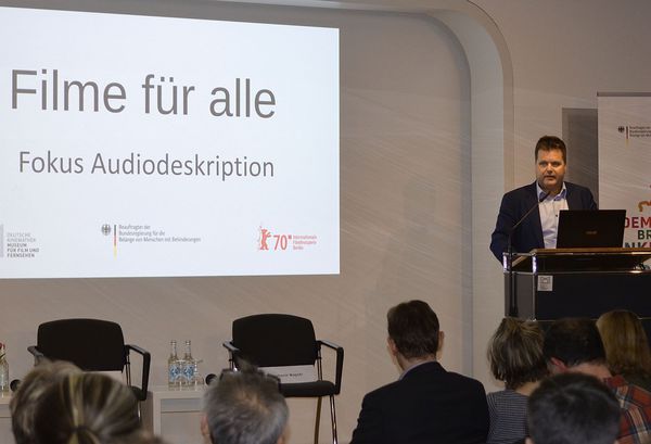 Jürgen Dusel steht am Rednerpult. Im linken Hintergrund sieht man die Projektion „Filme für alle - Fokus Audiodeskription“.