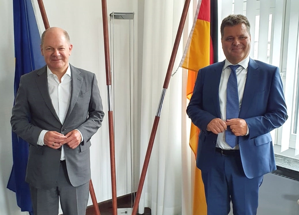 Vizekanzler Olaf Scholz und Jürgen Dusel stehen mit Abstand nebeneinander und lächeln. Im Hintergrund Flaggen.