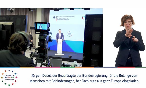 Man sieht ein Standbild aus einem Video. Jürgen Dusel ist sprechend zu erkennen, während er von Kameras gefilmt wird. Auf der rechten Bildseite wird in Gebärden übersetzt. Der untere Teil des Bildes wird für die Schriftdolmetschung genutzt.
