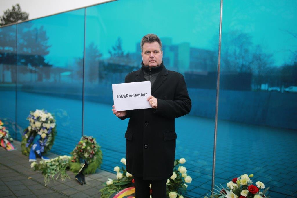 Jürgen Dusel steht vor der Blauen Wand und hält ein Schild mit Aufschrift #WeRemember.