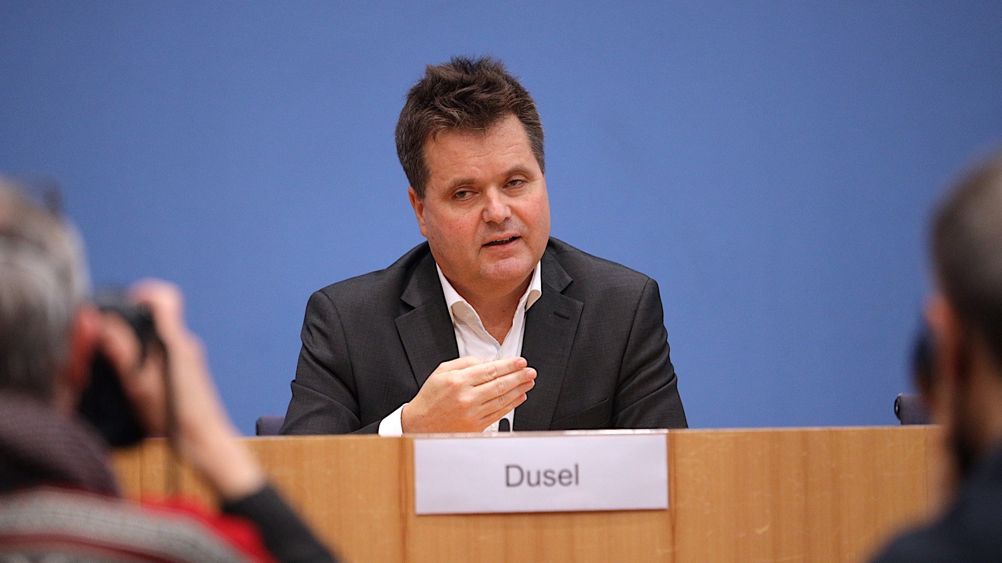 Jürgen Dusel an einem Pult, hinter ihm die blaue Wand der Bundespressekonferenz. Er spricht. Im Vordergrund unscharf Fotografen.
