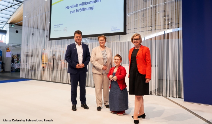 4 Personen (Dusel, Luczak-Schwarz, Fischer, Wirtz) stehen vor einem Vorhang und lächeln in die Kamera. Über ihnen ein Monitor mit der Aufschrift: Herzlich Willkommen zur Eröffnung!