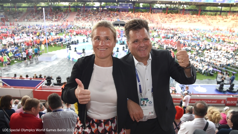 Jürgen Dusel und Verena Bentele stehen auf einer Zuschauertribüne. Sie lächeln in die Kamera und zeigen beide das Daumen hoch-Handzeichen. Hinter ihnen das Stadion voller Menschen.