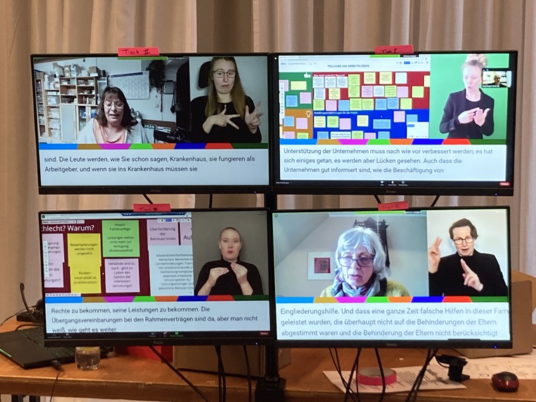 Foto von vier Bildschirmen, auf denen Personen, Wände mit Zetteln zu sehen ist. Außerdem Gebärdensprachdolmetscher*innen und Untertitel.