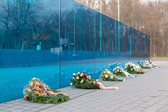 Vor einer blauen langen Glaswand liegen große Gedenkkränze mit Schleifen